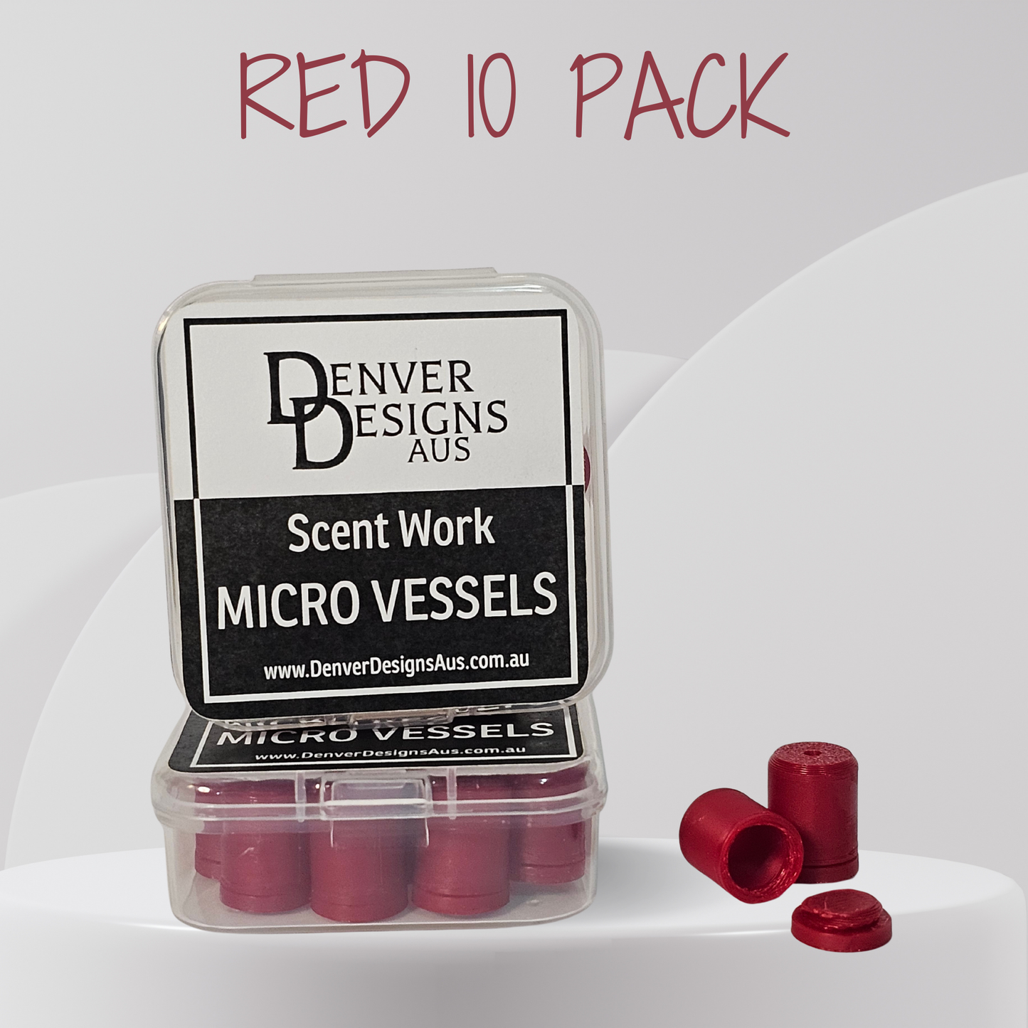 A clear plastic case containing small red cylindrical micro vessels, with three loose red vessels placed beside it on a white studio background. The lid label reads 'Denver Designs Aus' and 'Scent Work Micro Vessels.