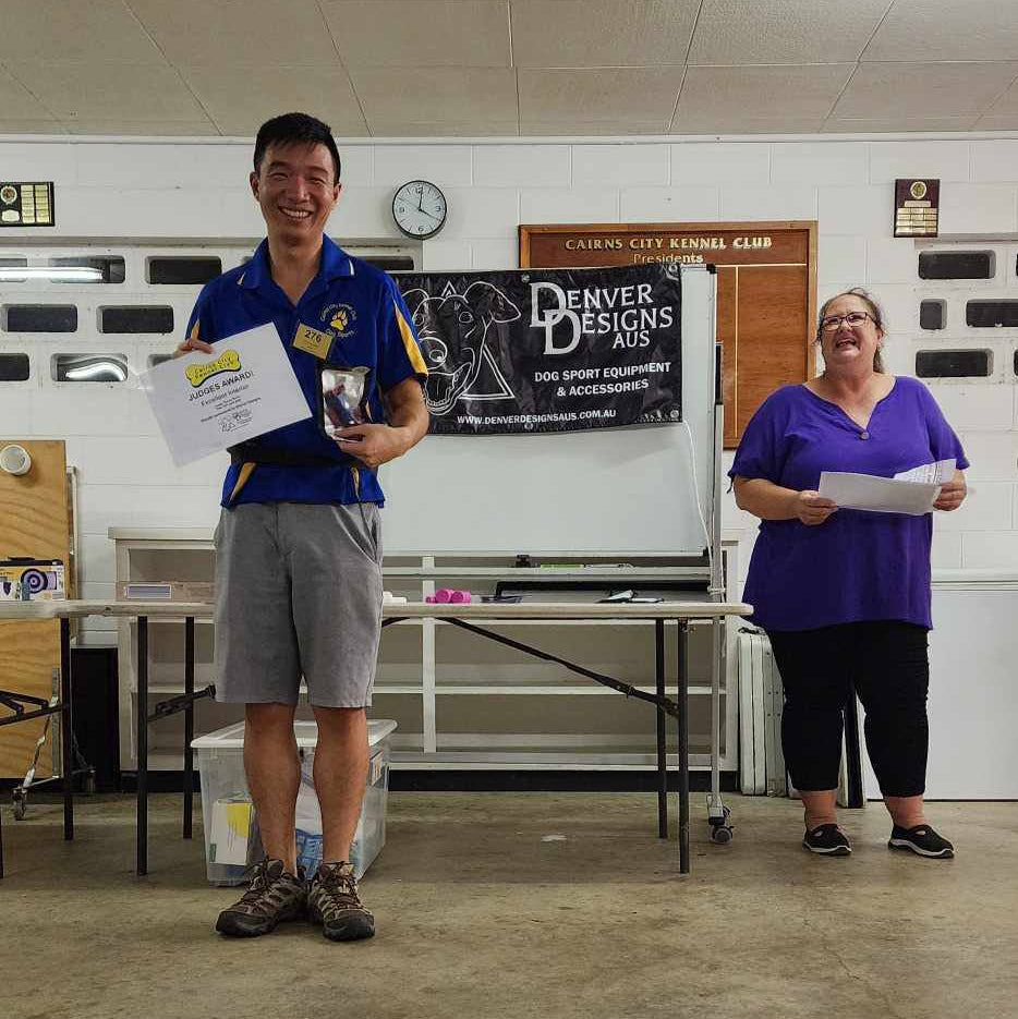 Two people standing in a room with a 'Denver Designs Aus' banner on the wall.