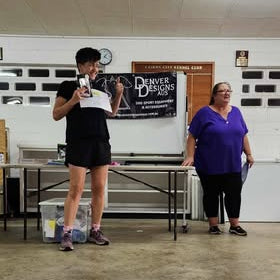 Two people standing in a room with a table and chairs, with a banner in the background.