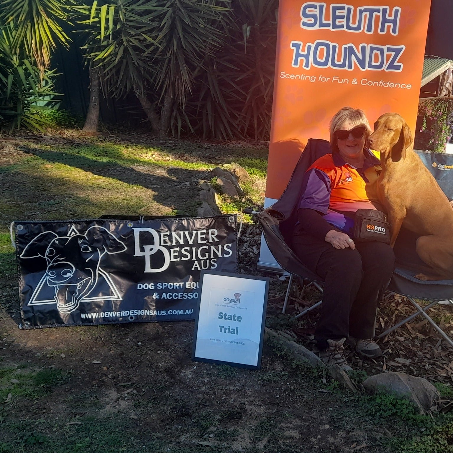 Woman with a dog sitting on a chair next to promotional banners for Denver Designs Aus and SLEUTH HOUNDZ.