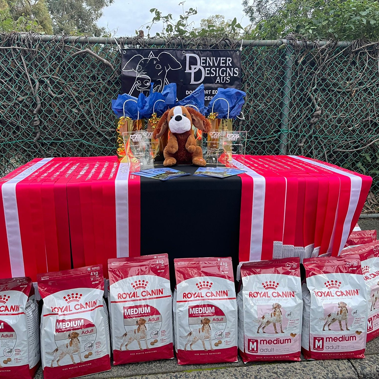 Table with Royal Canin dog food packages and Denver Designs plush toys outdoors.