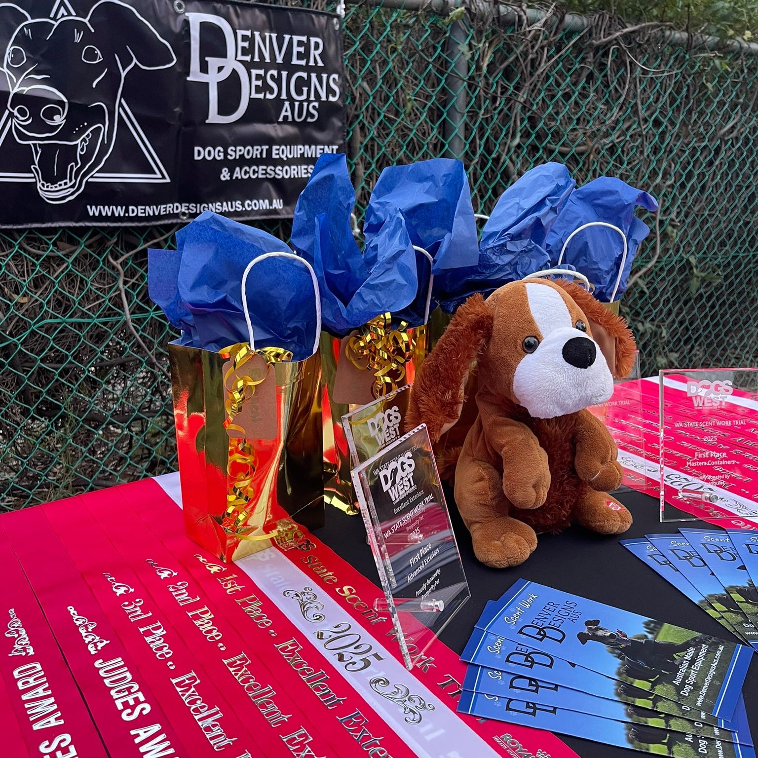 Dog show awards and accessories on a table with 'Denver Designs Aus' banner in the background
