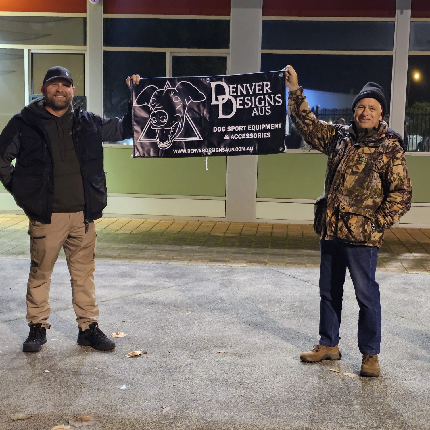 Two people holding a Denver Designs AUS sign in front of a store.