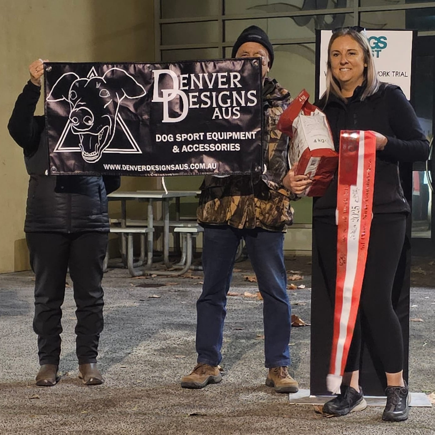 Three people holding a Denver Designs AUS sign and dog sport equipment.