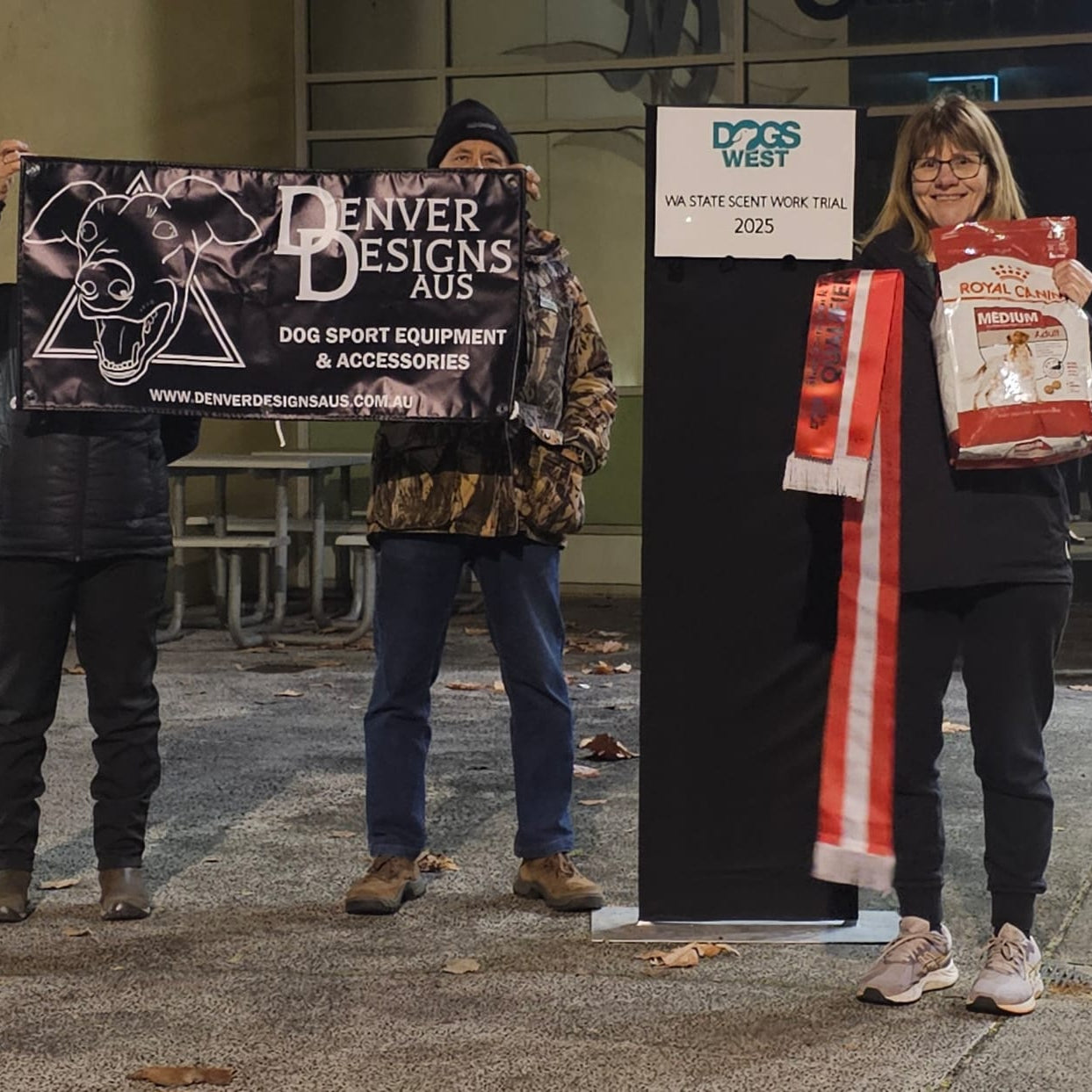 Two people holding a Denver Designs AUS sign and a woman with a red and white bag in an outdoor setting.