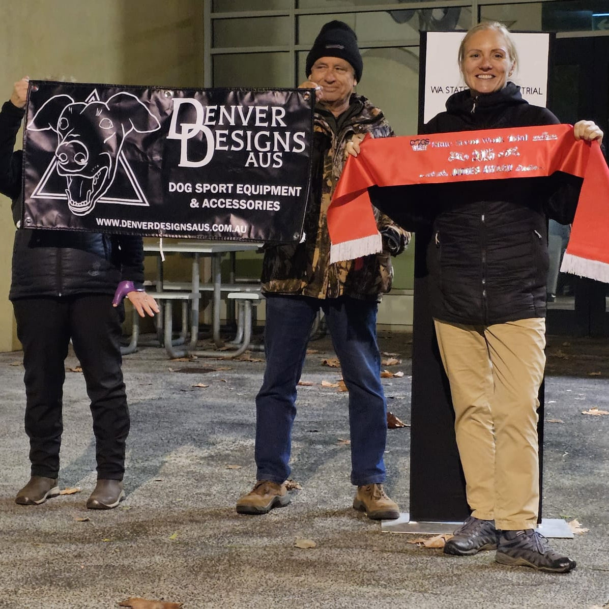 Three people holding up promotional materials for Denver Designs Aus in an outdoor setting.