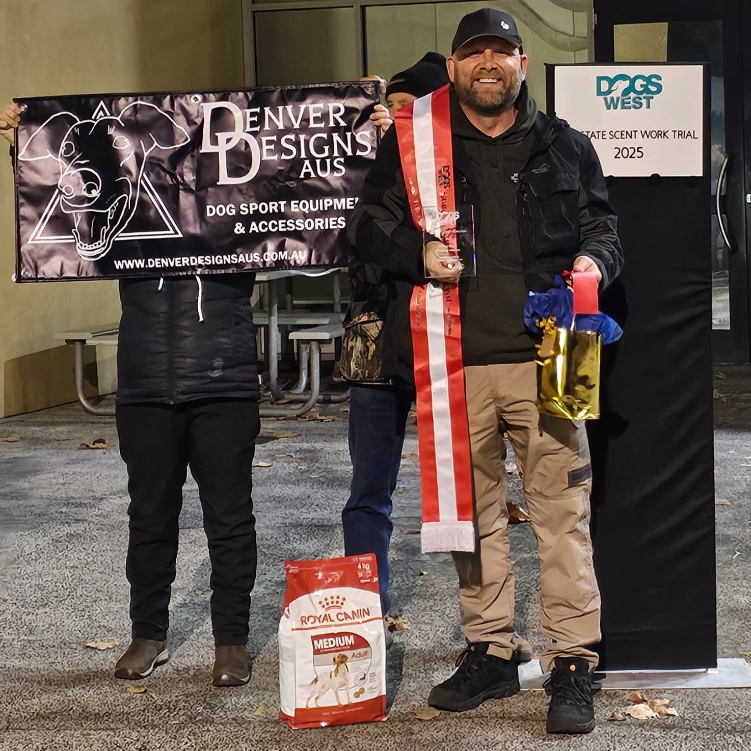 Person holding a Denver Designs AUS banner and a dog show ribbon, with a dog food bag and other items.