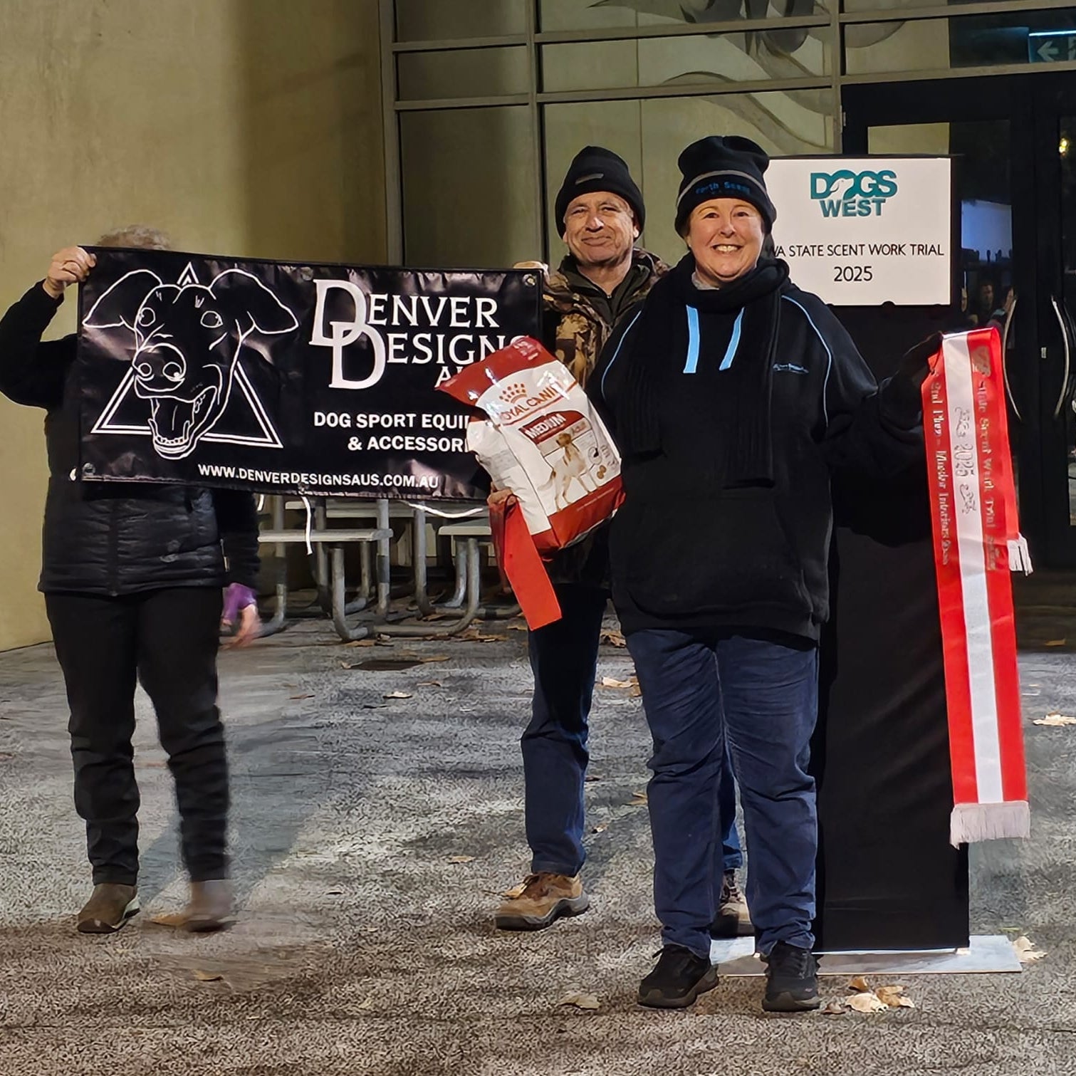 Three people standing outdoors holding signs and banners, with one person holding a bag of dog food.