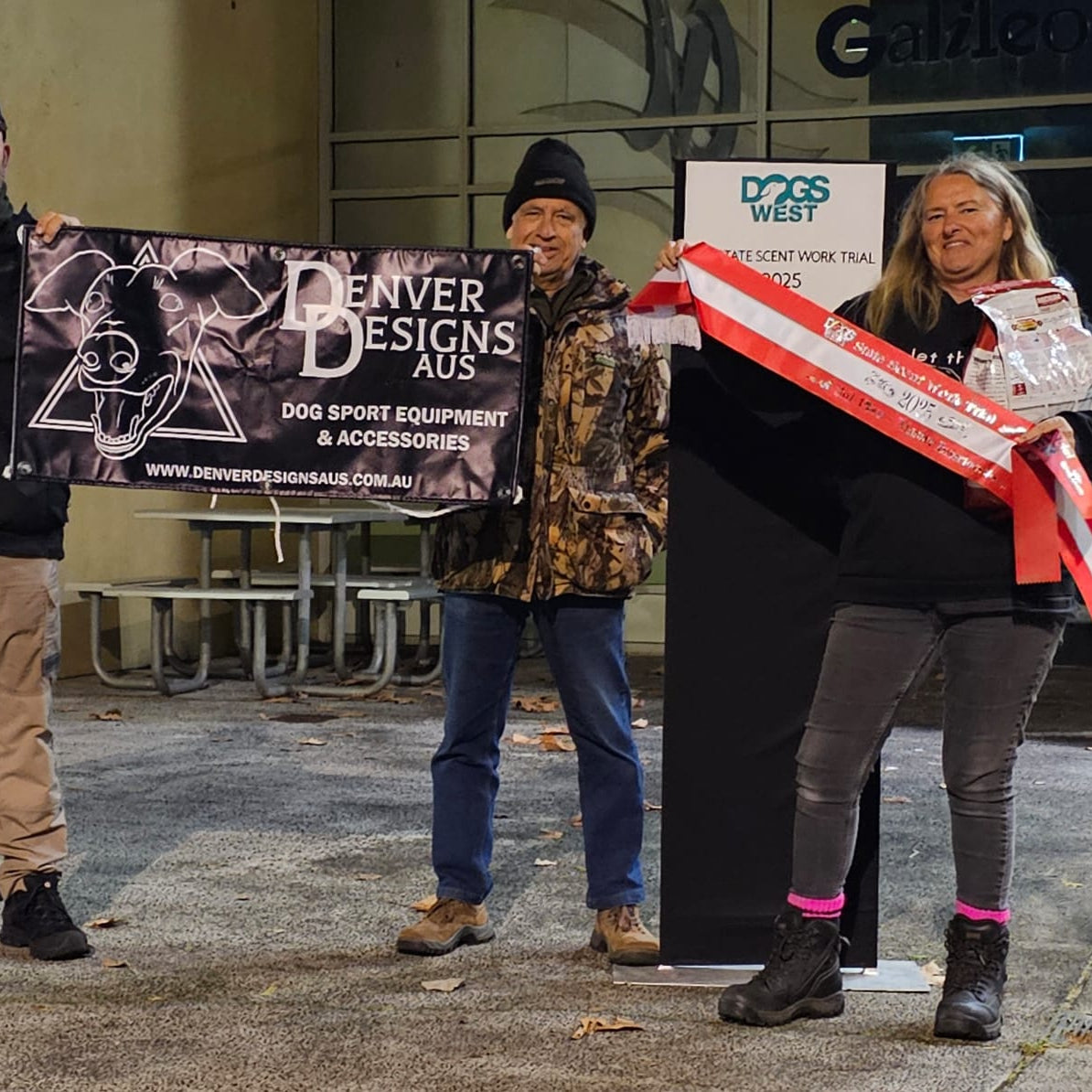 Three people holding promotional materials in front of a building with 'Denver Designs Aus' branding.