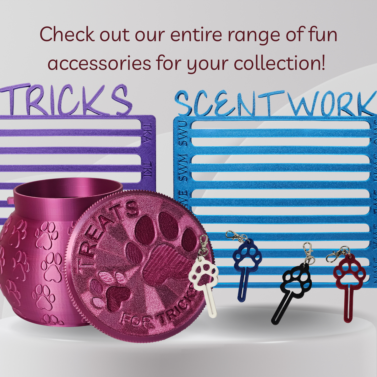 A promotional product shot showing a metallic pink cylindrical treat jar and matching round lid with embossed paw prints and the words 'TREATS FOR TRICKS,' alongside several key-shaped accessories in blue, black, white, and red. Large text reads 'Check out our entire range of fun accessories for your collection!' with 'TRICKS' and 'SCENTWORK' above the items.