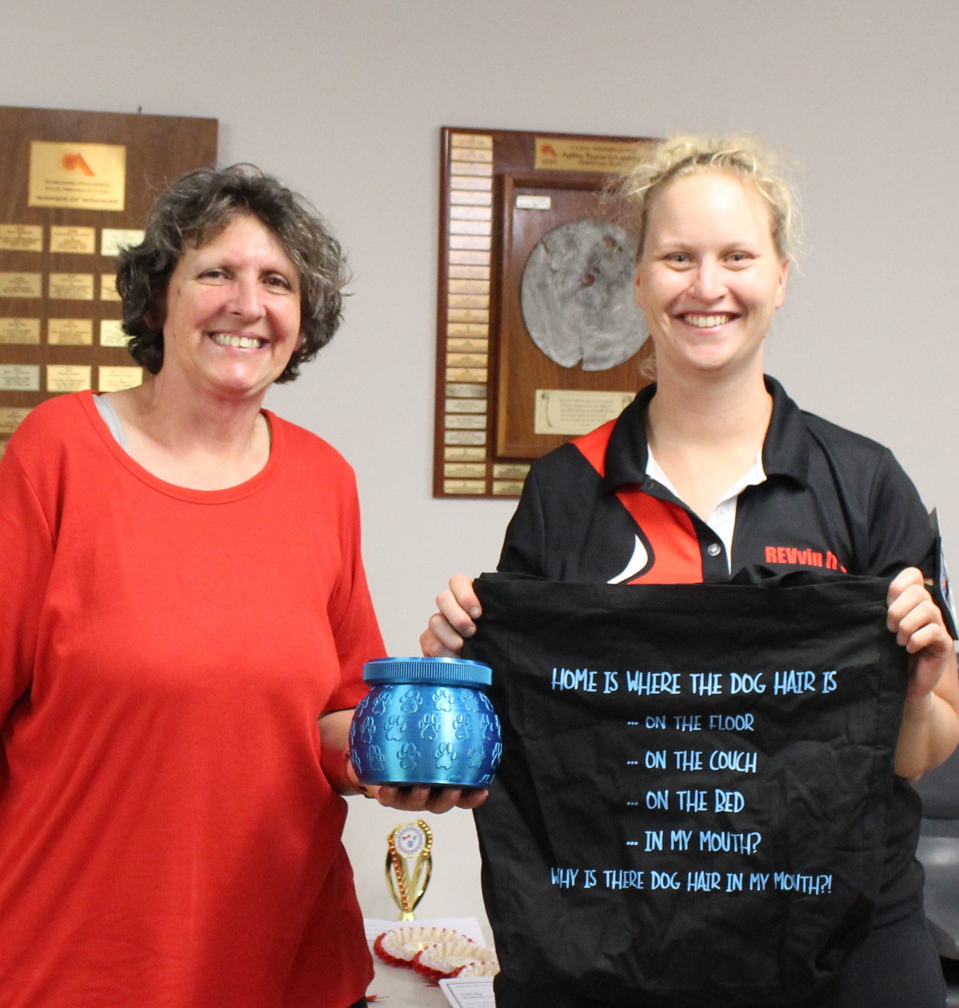 Two women holding a blue container and a black tote bag with humorous text in an indoor setting.
