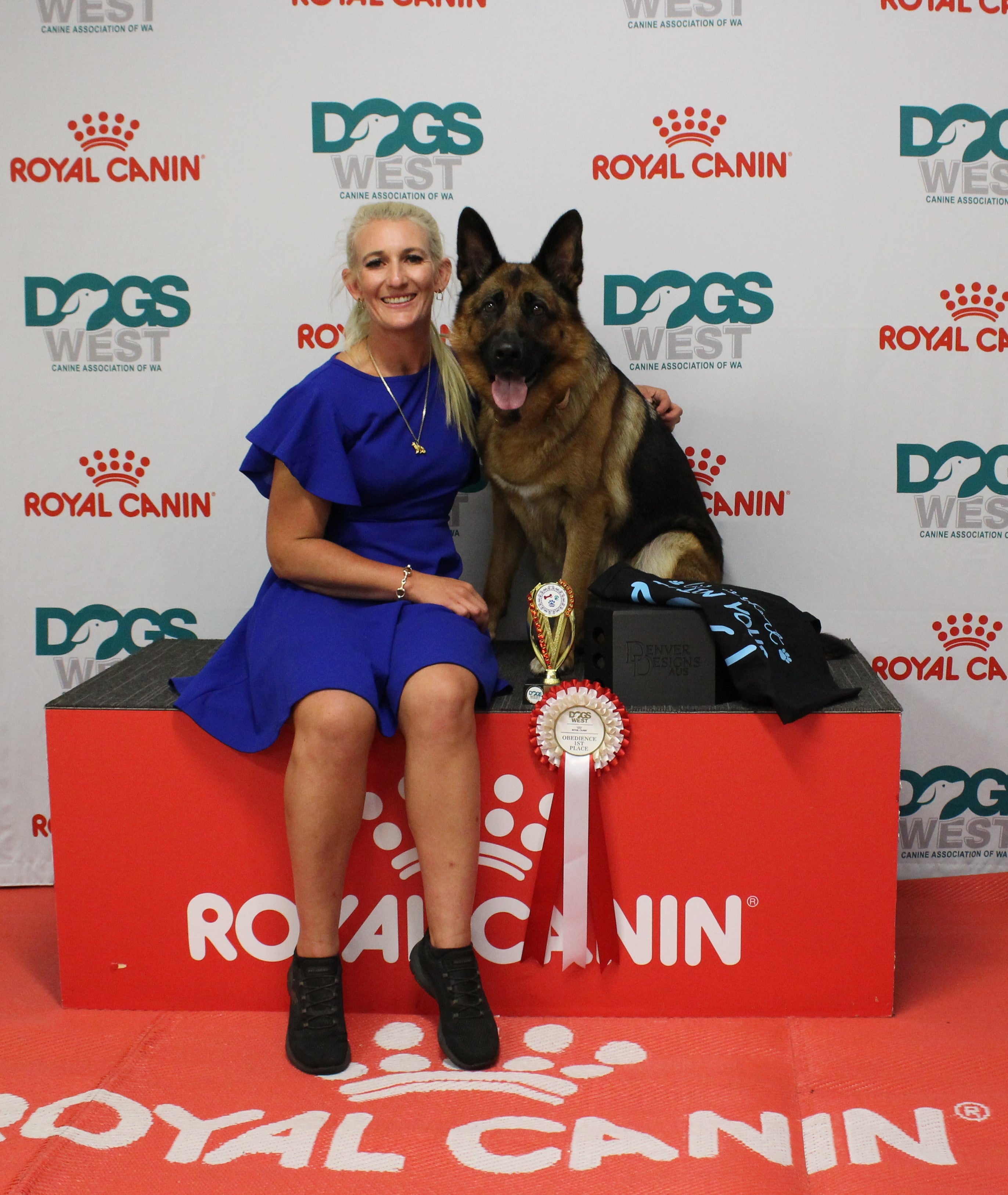 Woman in a blue dress sitting on a red platform with a German Shepherd dog, both holding awards, against a Royal Canin branded backdrop.