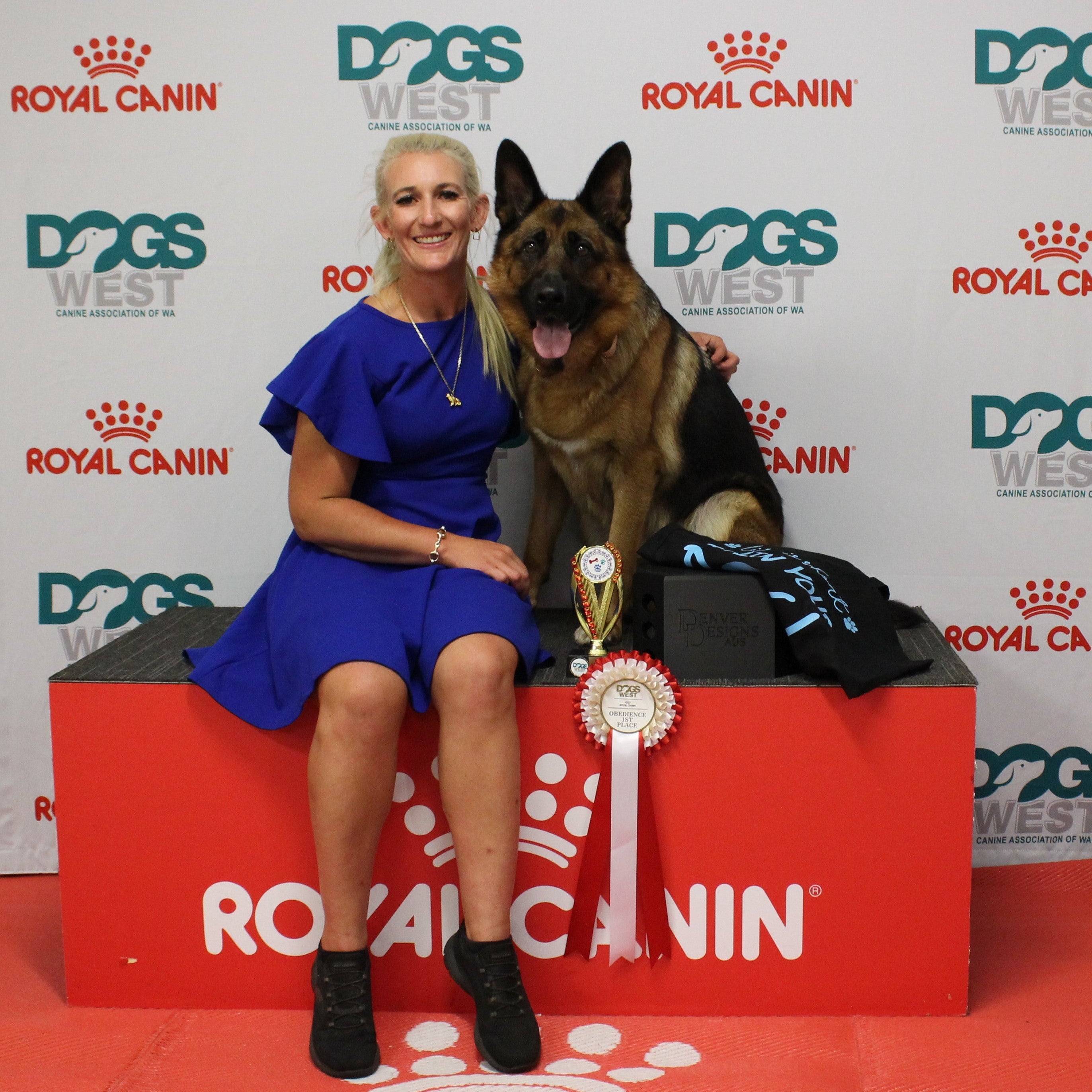 Woman in a blue dress sitting on a red platform with a German Shepherd dog, both holding awards, against a Royal Canin branded backdrop.